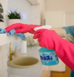 Close up of hands wearing rubber gloves cleaning a shelf with a cloth and spray bottle.